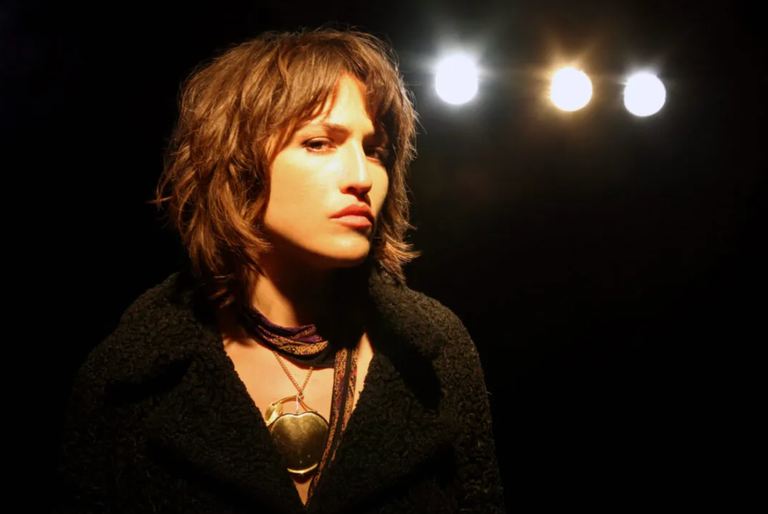 Portrait of musician Joan As Police Woman under stage lights wearing a dark coat and gold heart necklace.